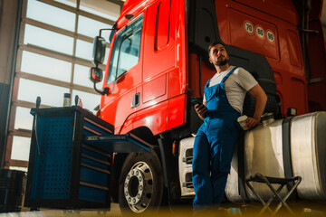 A mechanic in blue overalls leans against a red truck in a garage, holding a phone and a coffee...