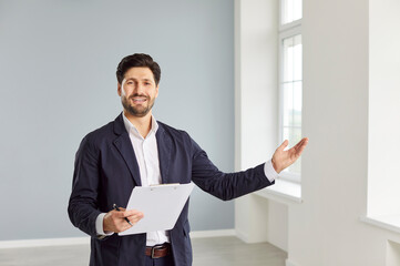 Happy smiling realtor or real estate agent showing a client a new apartment for purchase or rent. The agent standing in light room and holding clipboard in his hand. Buying and selling housing concept