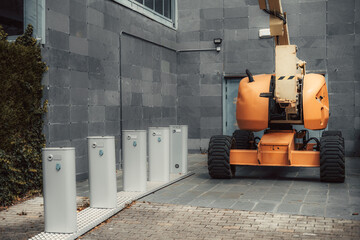 An industrial orange cherry picker parked beside a gray, tiled wall with ivy on the left side. In the foreground, five white utility boxes are aligned on a textured pathway