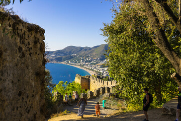 Fototapeta premium Alanya castle top view on the mountain with coast ferry boat on blue sea . Beautiful cleopatra beach Alanya Turkey landscape travel landmark.