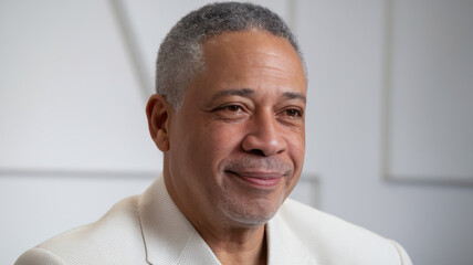 A middle-aged African American man with gray hair wears a stylish light-colored blazer, smiling warmly against a soft, neutral backdrop, exuding confidence and approachability.
