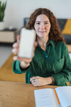 Young woman holding up a smartphone with a blank screen in a relaxed home office environment