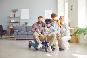 Happy joyful family, parents and excited funny little child sons at home wearing super hero fancy costume riding small tricycle, carefree two generations family having fun in new spacious living room