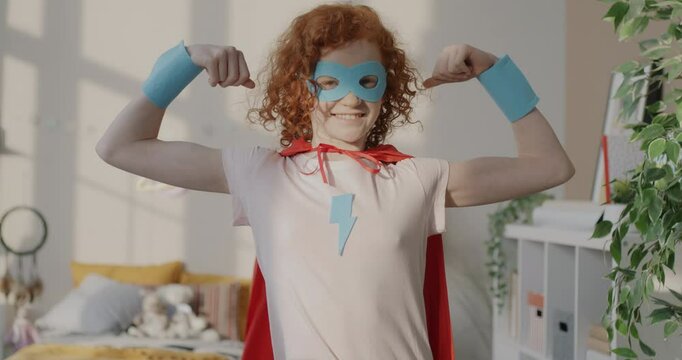 Slow motion portrait of funny little girl in superhero costume showing biceps muscles smiling looking at camera indoors at home. Childhood and fun concept.