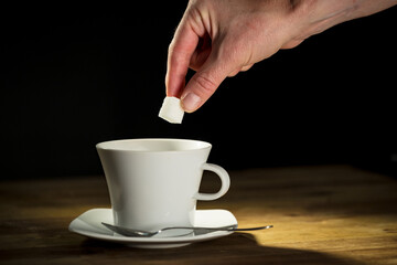 Male hand putting sugar cubes into a white coffee cup and stirring the drink with a teaspoon, close...
