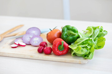 Closeup salad ingredients, including red and green peppers, tomatoes, radishes, and lettuce, arranged on a wooden cutting board