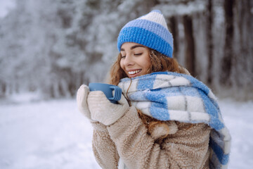 Beautiful woman is drinking a hot drink from a cup in the winter in nature. Winter forest. Travel, tourism, nature, active lifestyle.