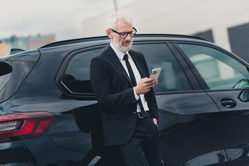 Mature businessman in formal suit checking smartphone near car on city street