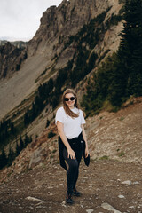 Naklejka premium The photo depicts a smiling woman standing on a mountain trail with a backdrop of majestic cliffs and forests.