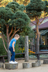 A little boy is jumping on a stump obstacle course in the yard, with a beautiful decorative pine tree in the background. Vertical photo