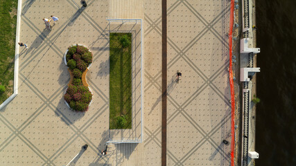 People walking and cycling on paved embankment near river in city park, aerial view