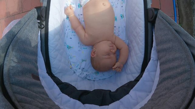 A infant in a nappy lying in a pram cradle. Top view. Close-up. The pram is covered with grey fabric on the outside and quilted with white fabric on the inside. A summer walk with a baby