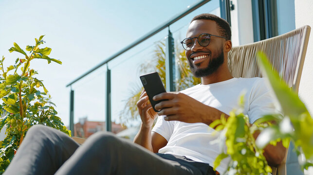 Modern outdoor relaxation: Smiling man seated on a comfortable chair on a balcony or terrace, engaging with his phone on a bright day. Perfect for illustrating enjoyment of outdoor