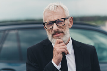 Mature businessman standing confidently outdoors near car, wearing formal suit and glasses, portraying thoughtful image