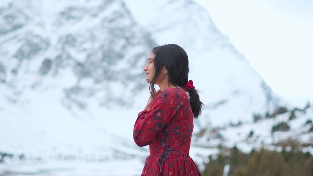 Indian woman wearing red dress and looking at snow covered mountains near Jispa in Lahaul, India. Girl enjoying fresh air in winter mountains. Travelling and exploring concept.