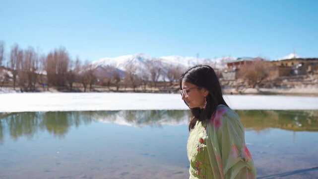 Beautiful Indian woman walking and half frozen Nako lake in background. Girl looking at frozen Nako lake and snowy Himalaya mountains in Nako Village, Himachal Pradesh, India. Winter travel concept