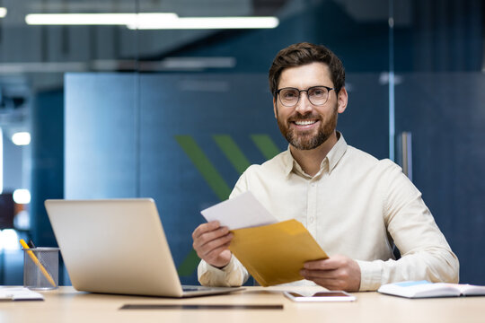 Portrait of a happy young businessman man who received good news and notification by letter, sitting in the office at the table, holding an envelope and looking at the camera