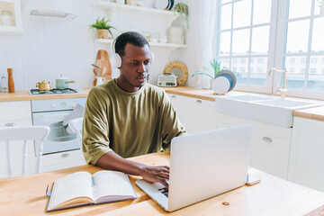 Young, focused black man wearing headphones sits at his kitchen table, working remotely on his...