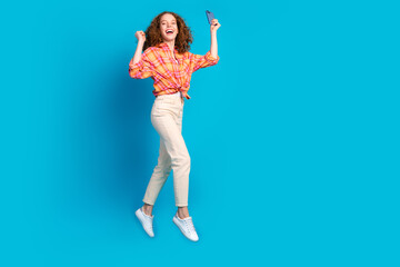 Joyful redhaired woman in plaid shirt celebrating success and gesturing against vibrant blue background, embodying happiness and positivity.