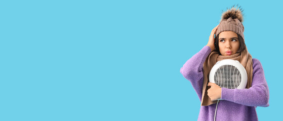 Fototapeta premium Beautiful young thoughtful woman in winter clothes with electric fan heater on blue background