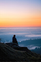 Obraz premium A person sits alone on a rocky hilltop, gazing over a misty forest landscape at sunset. Soft clouds fill the horizon, creating a peaceful and contemplative scene. Generative AI