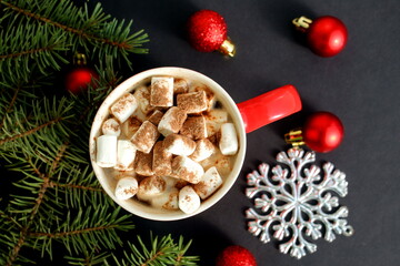 A red mug with cocoa and marshmallows stands on a black background with Christmas tree decorations.