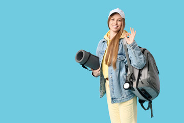 Smiling young woman with backpack and  camping mat showing OK gesture on blue background. Travel concept