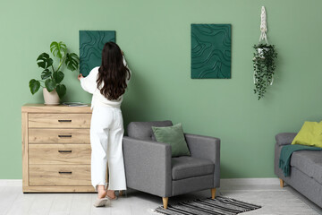 Young pretty woman hanging picture on green wall near wooden chest of drawers at home