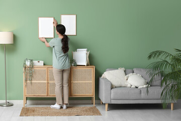 Young pretty woman hanging picture on green wall near wooden chest of drawers at home