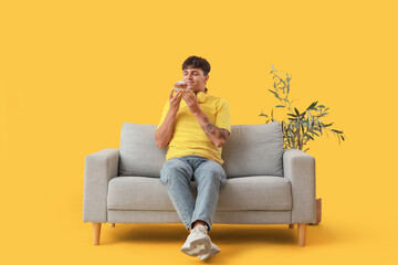 Obraz premium Young man with tasty muffin sitting on sofa against yellow background
