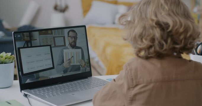 Over the shoulder view of child watching online lesson listening to teacher young man learning English with laptop and headphones at home. Childhood and education concept.