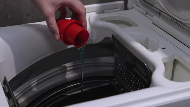 Woman pouring laundry gel into drum of top-loading washing machine, close-up of detergent dispenser