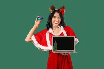 Pretty young woman in Santa costume with credit card and laptop on green background