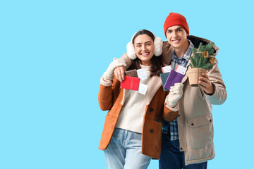 Cute young couple in warm clothes with small Christmas tree and passports on blue background. Winter vacation concept