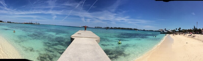 Panoramic view of a vibrant, tropical beach scene with turquoise waters, a concrete pier, and people enjoying various water activities.