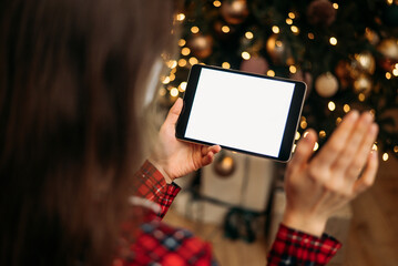 Woman holding tablet with blank screen in front of Christmas tree, festive bokeh lights in background, ready for holiday video call or virtual greeting.