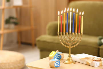 Menorah with burning candles, gift box and dreidels on table at home, closeup. Hanukkah celebration