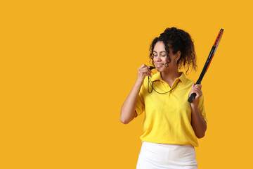 Female African-American tennis trainer with racket blowing in whistle on yellow background