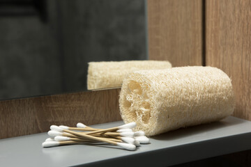 Close-up of a loofah sponge and wooden cotton swabs on a shelf. The blurred mirror and dark object in the background emphasize the natural and sustainable bathroom items