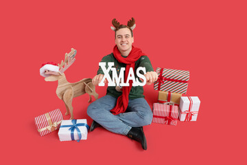 Young man with Christmas gifts, wooden reindeer and word XMAS on red background