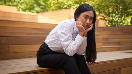 Fototapeta premium Sad Asian Korean Japanese woman sitting looking at camera student businesswoman bored upset tired lady lazy female business depressed burnout crisis pensive manager employer worker career girl outside