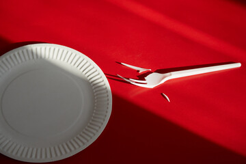 Disposable fork with broken tines next to a disposable plate in sunlight and shadow on a red...