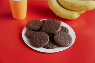 Chocolate cookies with coconut flakes on a white plate against a red background next to bananas and a disposable orange cup, chocolate cookies on a plate.