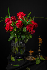 View of red roses with green leaves in a vase on a table with 2 lit candles, black background, vertical, with copy space