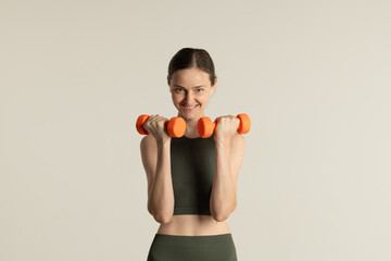 The woman holds orange dumbbells with a confident smile, highlighting her commitment to fitness in a bright, minimalistic environment dedicated to workout activities.