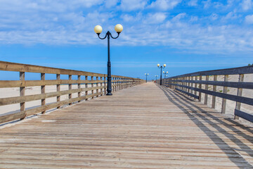 A serene wooden promenade with lampposts extends along the sea beach under a bright blue sky in the early afternoon