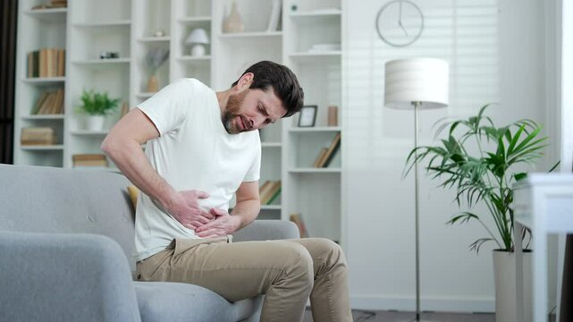 Upset handsome man feels stomach pain while sitting on sofa in living room at home. Sick bearded male has heartburn, gastritis, bloating or poisoning. Male suffers from spasms and constipation