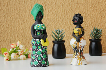 Two statuettes of black women wearing traditional green and yellow brazilian costumes. Orange color wall background. Vases of succulent plants and a bunch of rose buds on the composition.