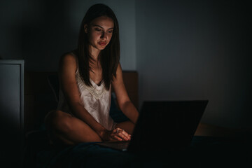 A young woman is sitting in a dimly lit room, deeply focused on her laptop. The night setting...