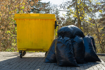 A striking yellow garbage can rests next to a heap of black garbage bags, set against a backdrop of colorful autumn leaves and clear skies, reflecting a quiet urban moment